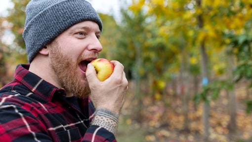 A man outdoors taking a bite of a fresh apple, wearing a knit hat and plaid shirt