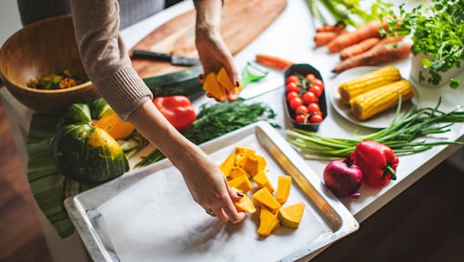 Hands preparing a colorful assortment of fresh vegetables on a kitchen counter, including squash, tomatoes, carrots, corn, and leafy greens.