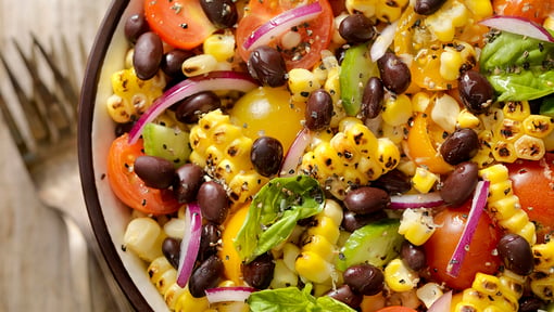 Bowl of a colorful plant‑based meal with black beans, corn, tomatoes, avocado, red onion, and leafy greens.