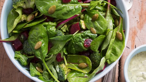 Bowl of leafy green salad with spinach, mixed greens, seeds, and vegetables, set on a wooden table.