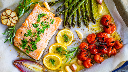 Baked salmon fillet served with asparagus, roasted tomatoes, sliced lemon, garlic, and herbs on a parchment‑lined baking sheet.