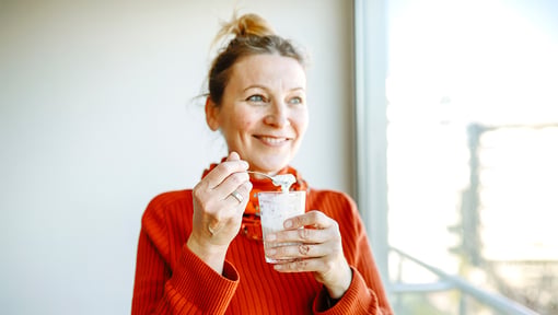 A woman holding a glass of yogurt topped oatmeal and spoon indoors near a window.
