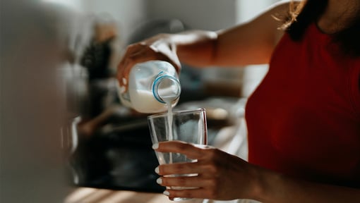 Hand pouring bottled milk into a clear glass in a kitchen setting.