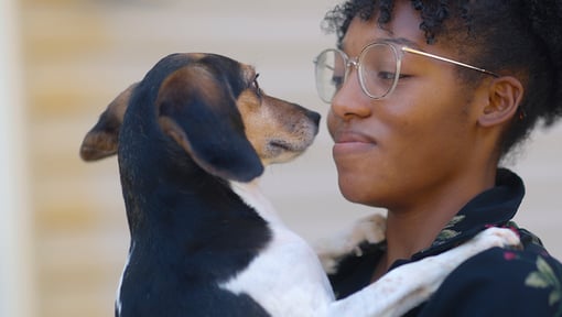 Close-up of a woman wearing glasses holding a small black-and-white dog at face level, with the dog looking toward the woman.