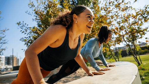 two women holding planks on a low wall during an outdoor workout session. 