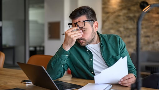 A man seated at a desk holding papers and rubbing the bridge of his nose, with an open laptop on the table.