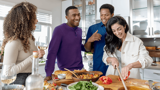 Group of people preparing food together in a bright kitchen, with one person chopping vegetables on a cutting board and others standing nearby.