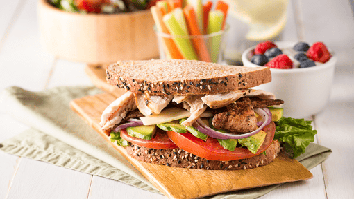 Healthy whole‑grain turkey sandwich layered with lettuce, tomato, red onion, and avocado, served on a wooden board with berries, celery, and carrot sticks.