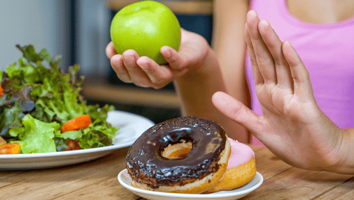 Person holding a green apple while raising a hand to decline two donuts on a plate.