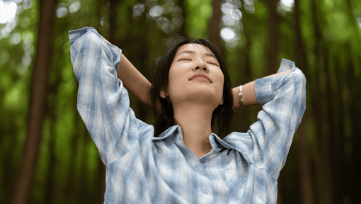 A woman enjoying a mindful moment in nature, her hands are behind her head with her eyes closed.