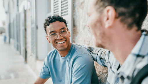 A man looks at a friend with a smile on his face as they sit outside on a bench.