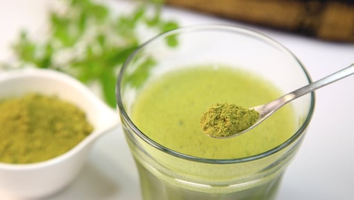 A close up of a glass filled with green drink, a spoon of Moringa superfood held over the glass about to be poured in.