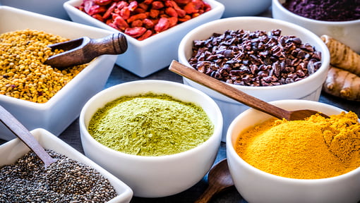 Bowls of colorful superfoods including turmeric powder, matcha, chia seeds, goji berries, bee pollen, cacao nibs, and fresh ginger.