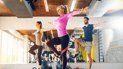 Three people in a fitness studio performing a standing balance exercise, each raising one knee while extending both arms out to the sides.