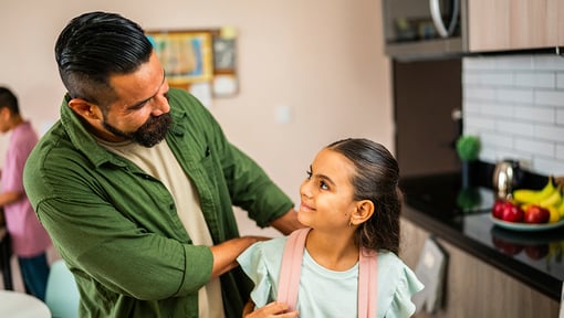 Adult and child standing in a kitchen near a counter, making eye contact while the adult adjusts the child’s backpack straps.