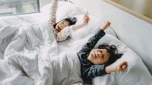 Two young children lying in a white bed, stretching their arms overhead as they wake up, surrounded by pillows and a white comforter in a softly lit bedroom.