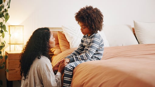 Adult kneeling beside a bed, holding hands with a child sitting on the edge in striped pajamas.