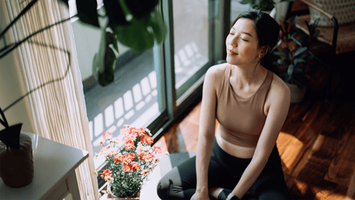 A woman sitting on a wooden floor near a sunlit window, wearing athletic clothing, with a potted plant and small flowers beside them