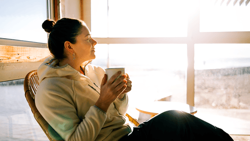 A woman sitting in a chair by a large sunlit window, holding a mug with both hands
