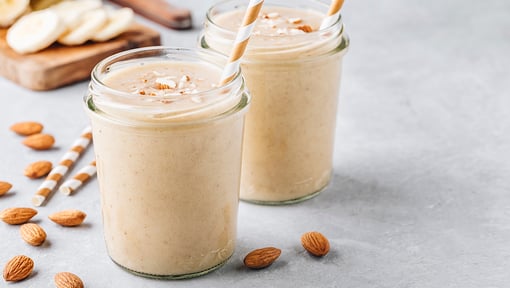 Two jars of banana almond smoothies with striped paper straws, surrounded by whole almonds and sliced bananas.