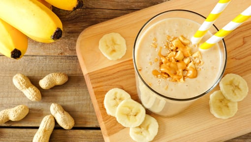 Banana smoothie topped with peanut butter and oats in a glass, surrounded by banana slices and peanuts on a wooden table.