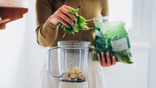 Person adding fresh spinach to a blender containing banana slices while preparing a green smoothie in a bright kitchen.