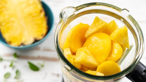 Fresh pineapple chunks and mango pieces inside a blender pitcher, with a sliced pineapple visible in the background.