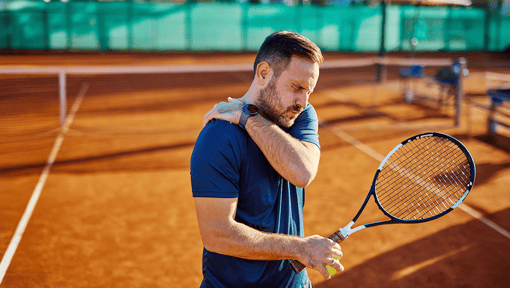 Person standing on a clay tennis court holding a tennis racket and touching one shoulder, suggesting shoulder discomfort or injury during play.