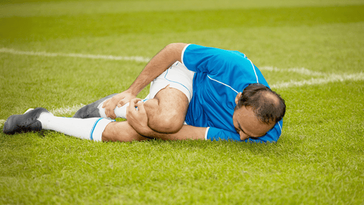 Man lying on a grassy sports field holding a bent knee, appearing to have a knee injury during athletic activity.
