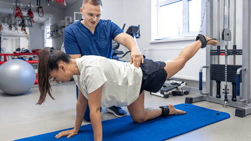 Physical therapist guiding a person through a rehabilitation exercise on a blue mat, with the person raising one leg while using hands and knees for support in a therapy clinic.