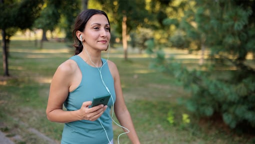 A woman walking and listen to something on her smart phone on a tree-lined outdoor path.