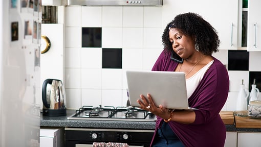 A woman multitasking using a laptop and smartphone while working in the kitchen.