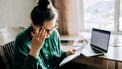 A woman sitting at a desk with a laptop, appearing overwhelmed by multiple tasks.