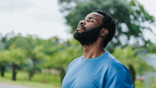 A man standing outdoors with his head turned up to the sky with his eyes closed, taking a mindful moment in nature to relax and recharge.