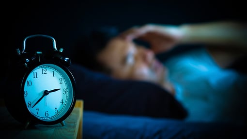 Alarm clock on a bedside table with a person lying awake in bed in the background.