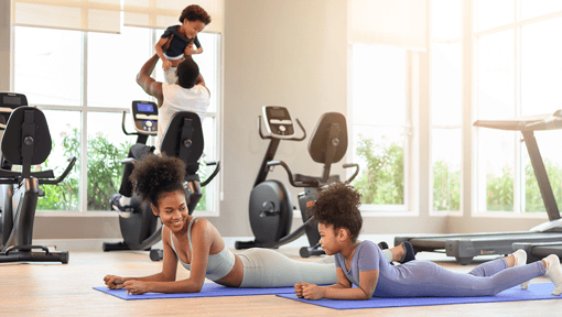 Family-friendly gym environment with two adults and two children staying active through floor exercises, cycling, and fitness activities indoors.