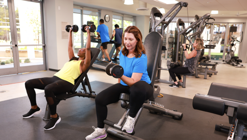 People participating in a strength-training workout at a fitness center, using dumbbells, benches, and resistance machines.
